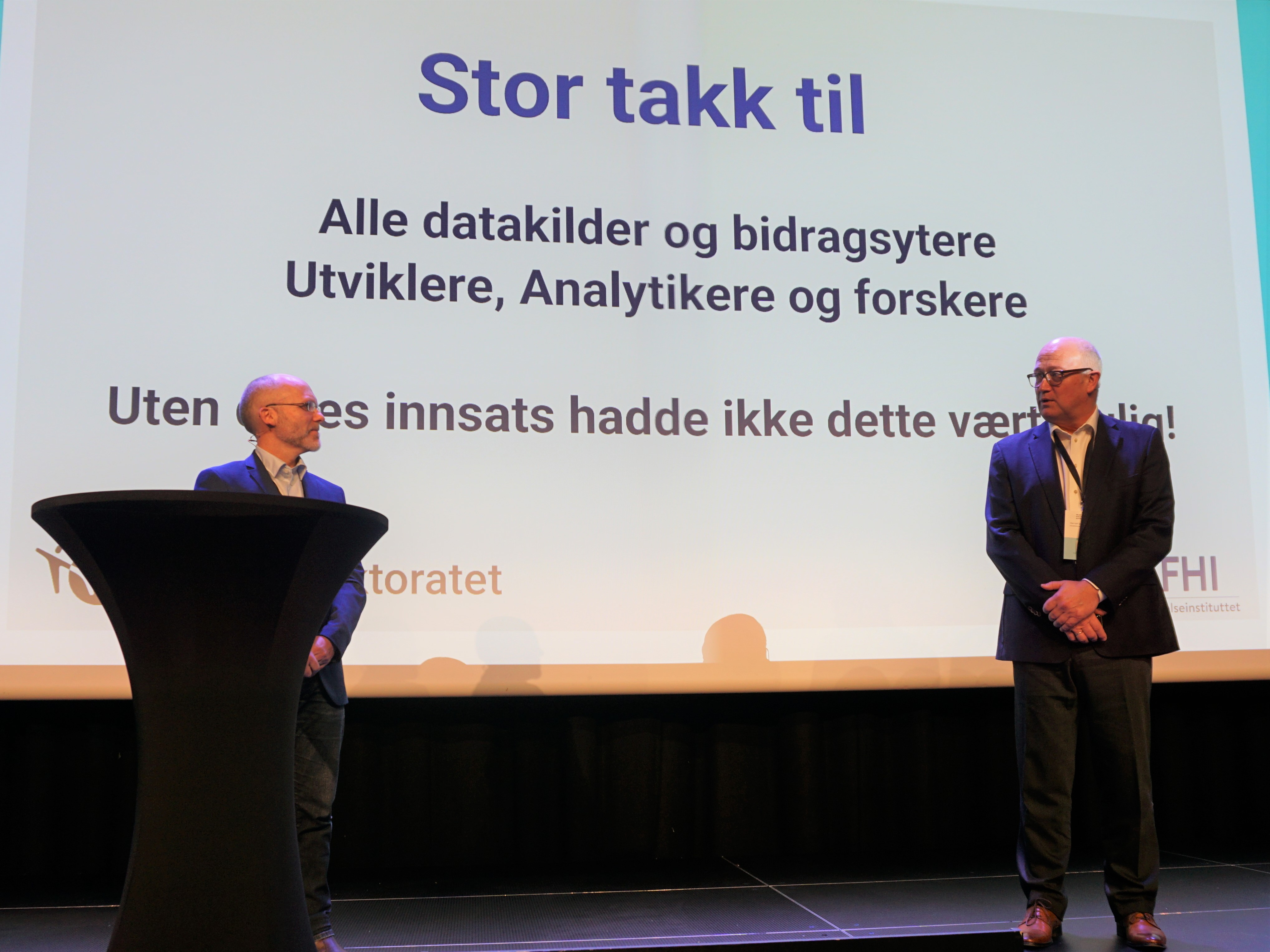 En mann og en kvinne står ved en talerstol med en stor skjerm bak seg