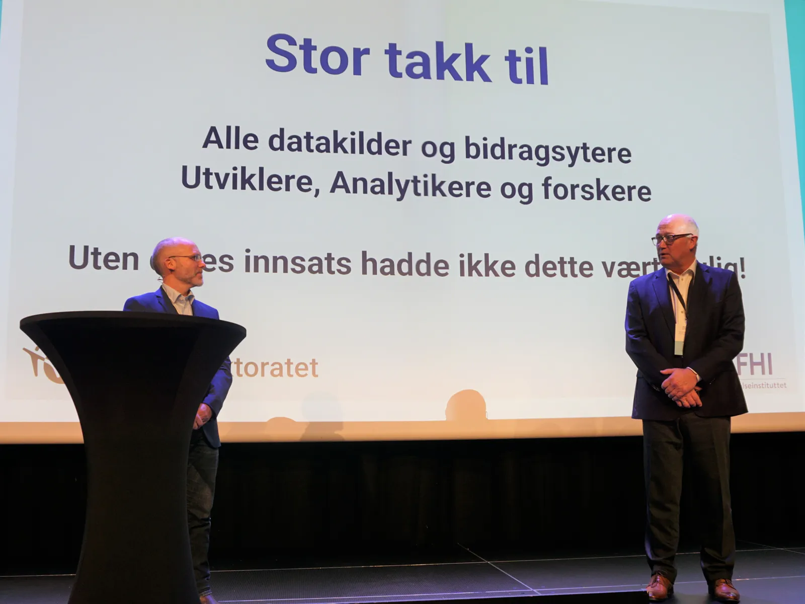 En mann og en kvinne står ved en talerstol med en stor skjerm bak seg