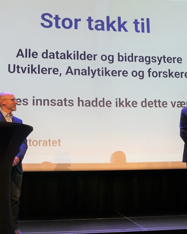 En mann og en kvinne står ved en talerstol med en stor skjerm bak seg